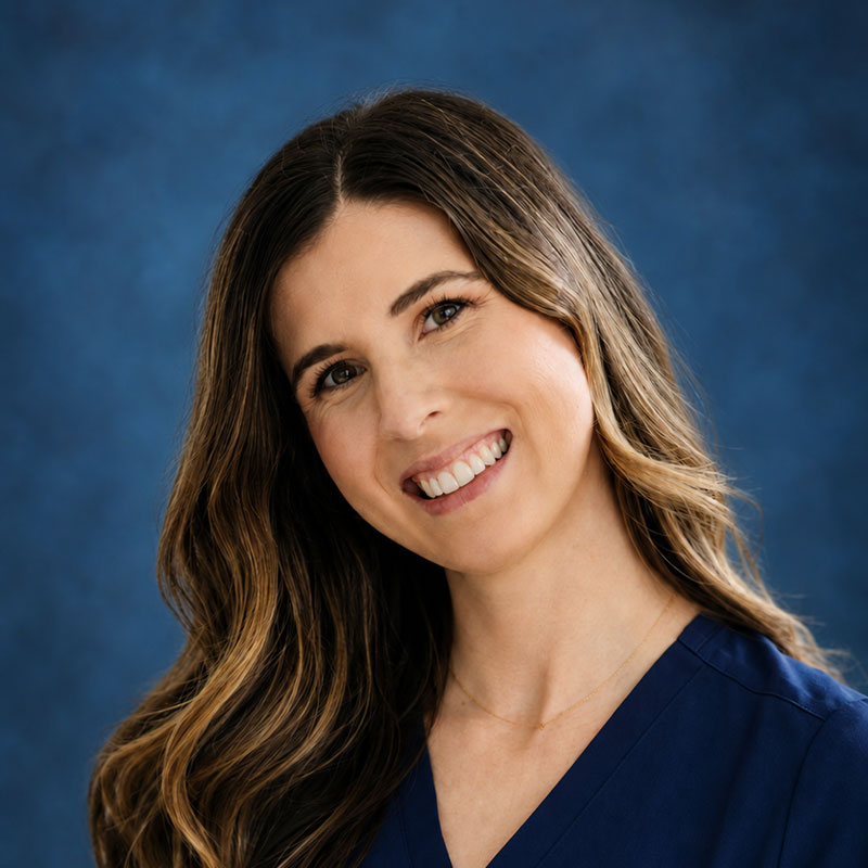 A woman with long brown hair, wearing a navy blue top, smiles against a blue textured background. Kailee Sardelli, BSN, RN, radiates warmth and professionalism in this vibrant setting.