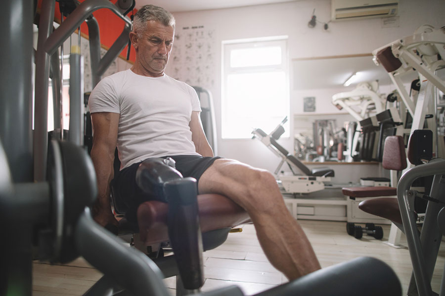 An older man with a prosthetic leg, managing knee osteoarthritis, uses a leg extension machine in a gym, wearing a white t-shirt and black shorts, focused on his workout. Fitness equipment surrounds him in the well-lit room as he maintains his routine—an inspiring moment worthy of any journal.