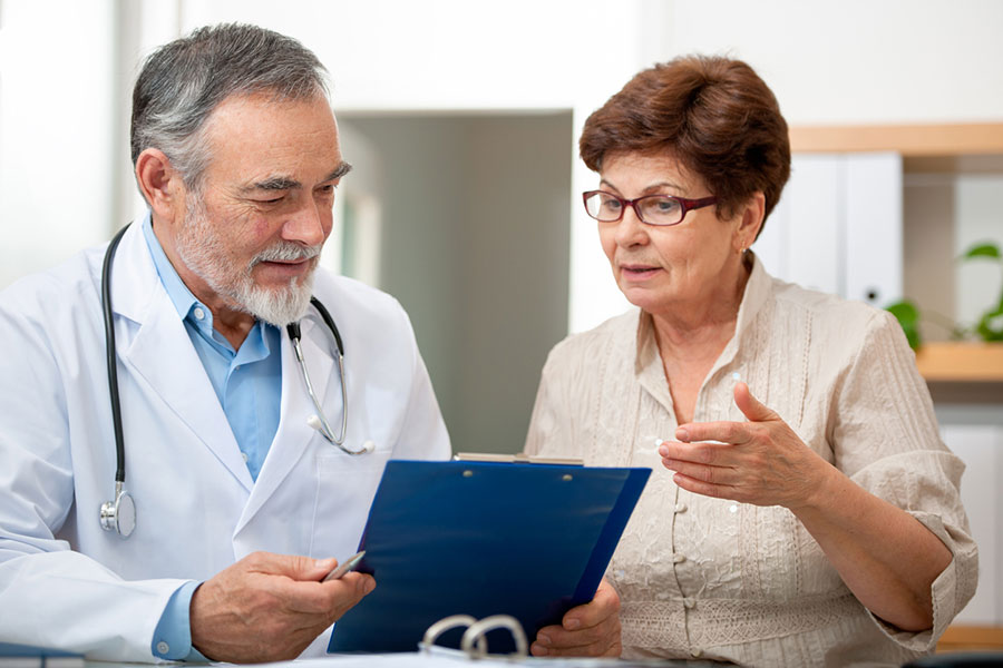 An older woman speaks with a male doctor holding a clipboard. As they discuss medical information in a well-lit office setting, the conversation touches on regenerative medicine—what regenerative medicine means and how it could impact her treatment options.