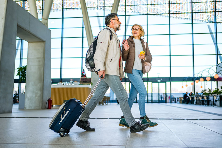 A man and woman, taking a natural approach to travel, walk through a modern airport terminal with large windows. The man pulls a suitcase, and both appear to be talking and smiling. They wear casual jackets, carry backpacks, and look refreshed as they make their way through the spacious terminal.
