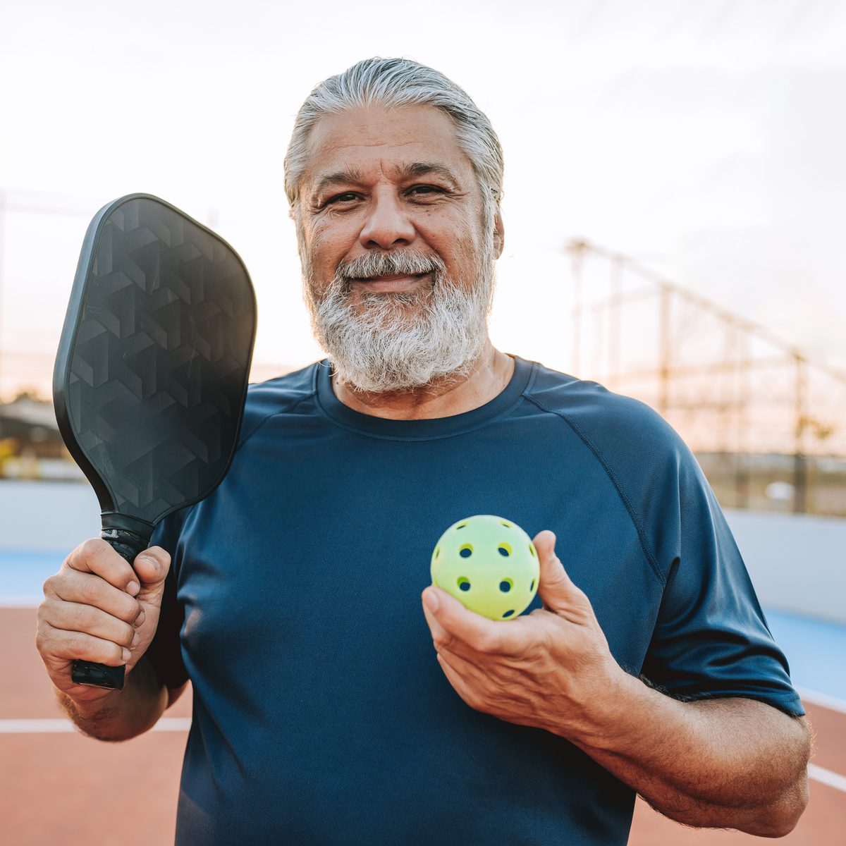An older man with a grey beard holding a pickleball paddle and a pickleball on a court.
