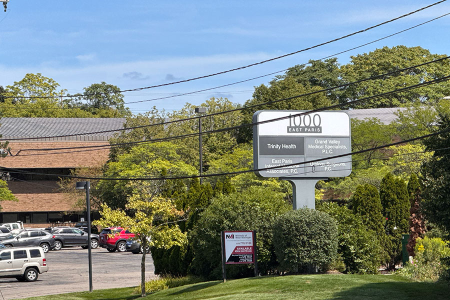 A sign for 1000 East Paris listing Trinity Health and other medical offices, with a smaller NAI real estate sign in front of a parking lot and workspace building in Grand Rapids.