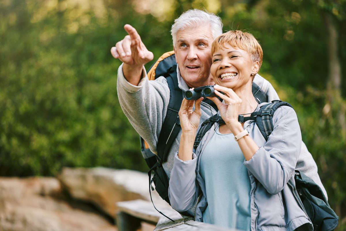 An older man points into the distance while an older woman looks through binoculars.