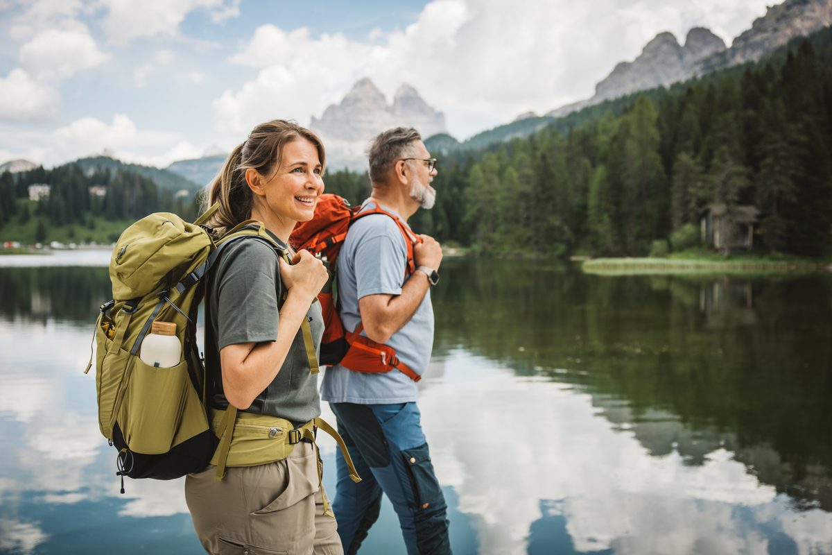A smiling woman and a man wearing hiking backpacks stand by a mountain lake.