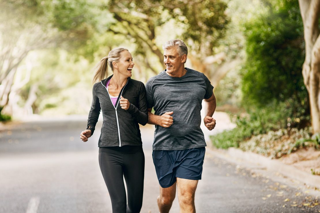 An older man and woman laughing while jogging together on a tree-lined road.