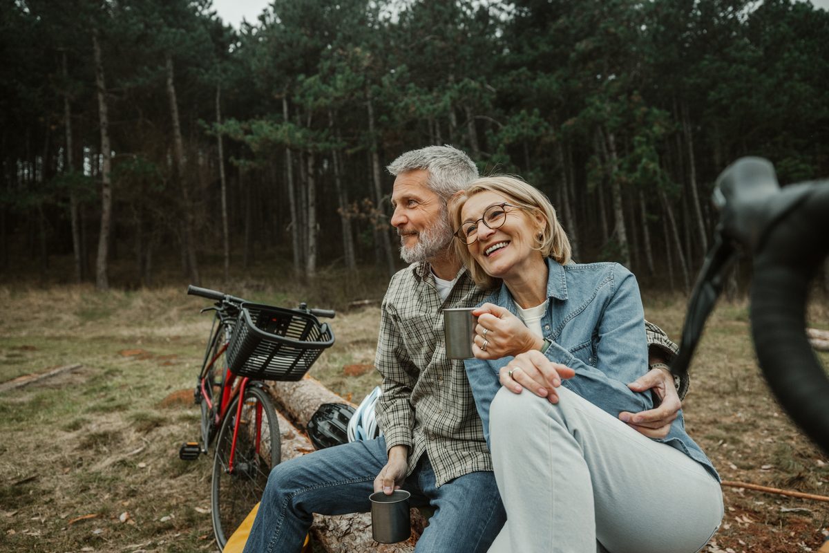 A middle-aged couple sits on a log in a forest, holding mugs and smiling, with a bicycle nearby.
