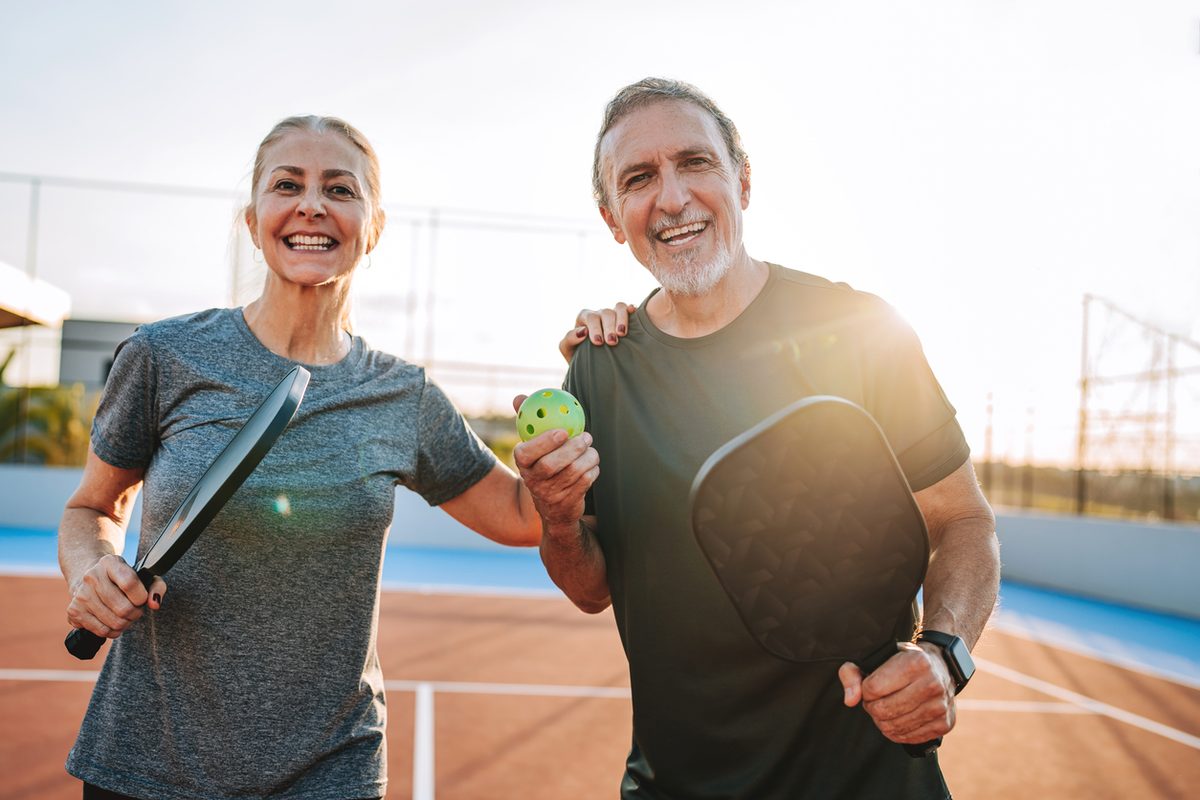 A smiling man and woman holding pickleball paddles and a ball on a court