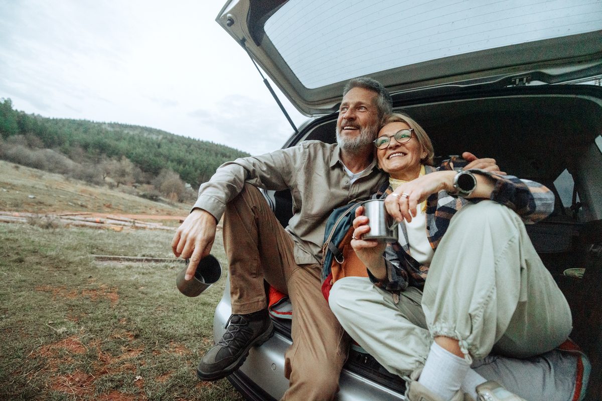 A smiling middle-aged couple sitting in the open trunk of a car, holding metal mugs while looking out at a hilly landscape.