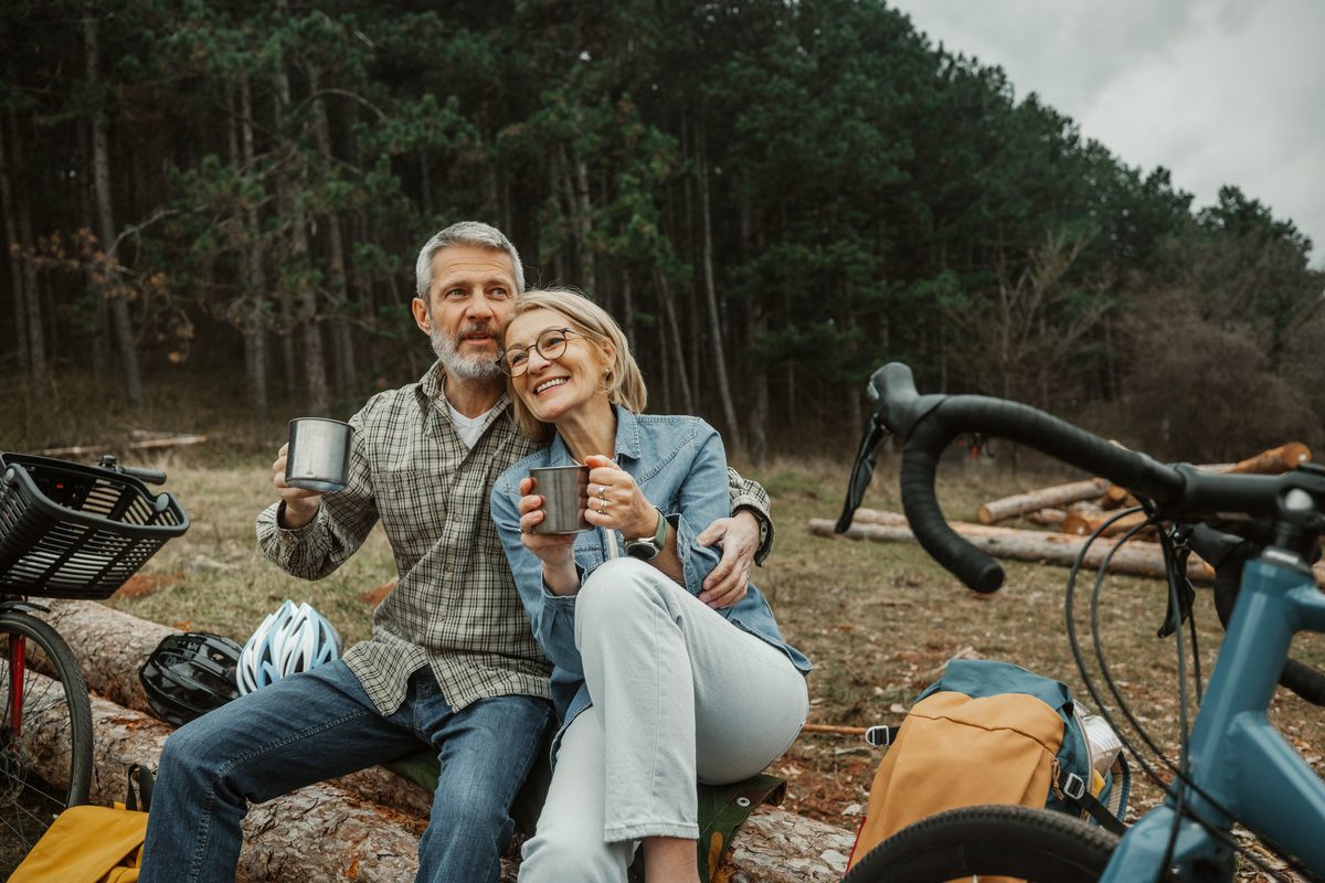 A smiling couple sits on a log in the woods holding metal camping mugs, with bicycles parked nearby.