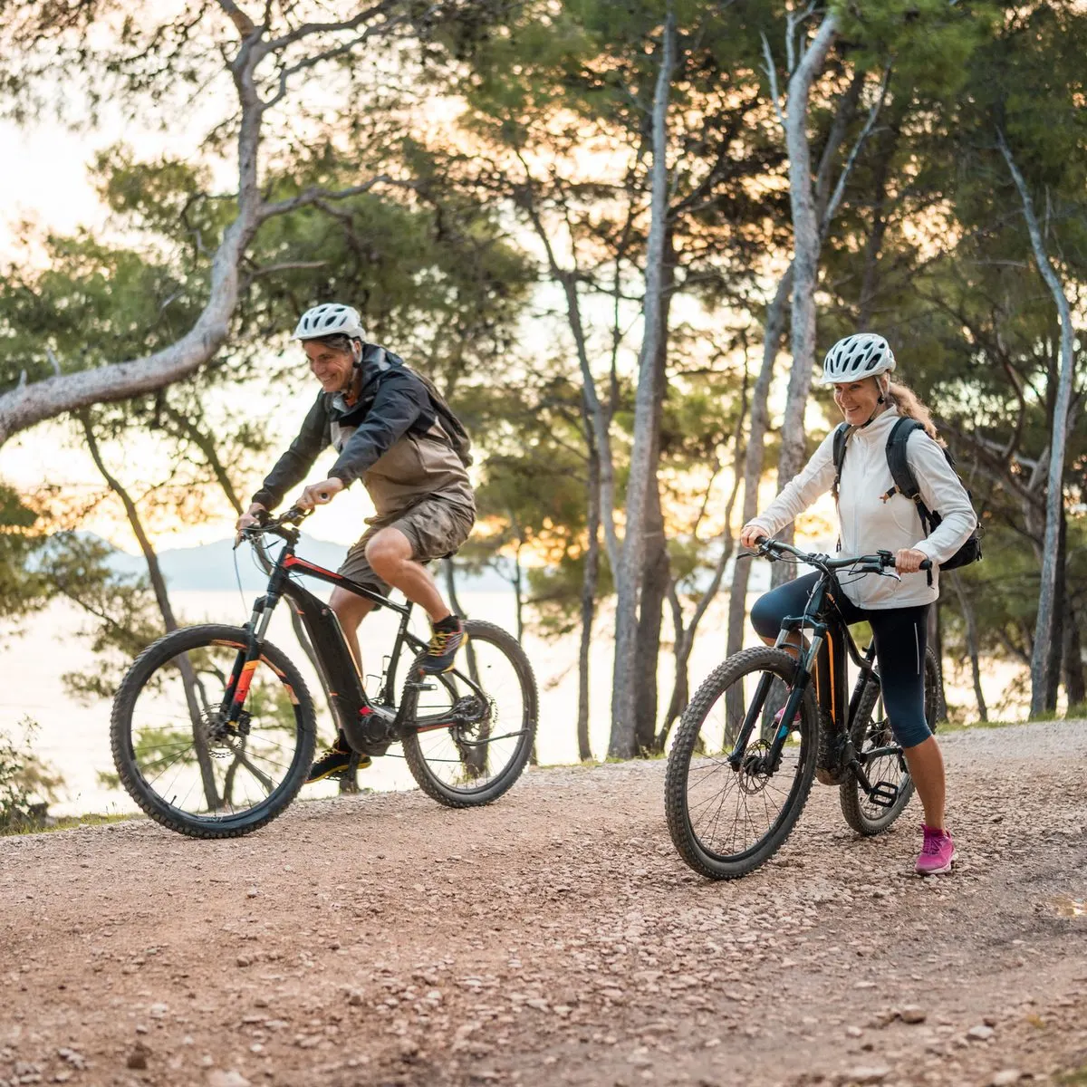 A man and woman wearing helmets riding electric mountain bikes on a dirt path in a wooded area near the water.