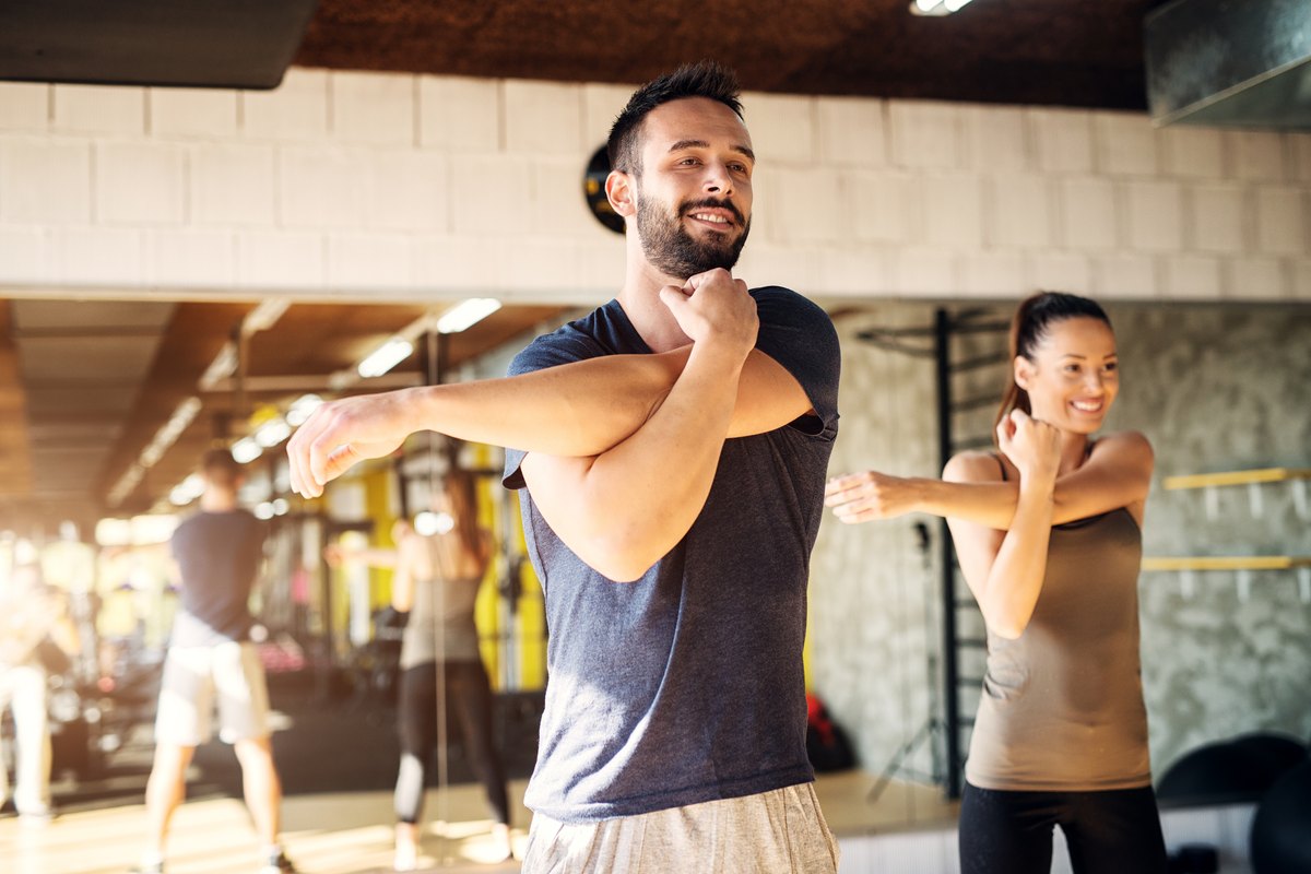 A man and a woman in a gym performing cross-body arm stretches