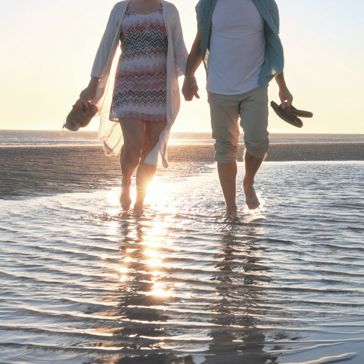 A couple holds hands while walking barefoot through shallow water on a beach at sunset.
