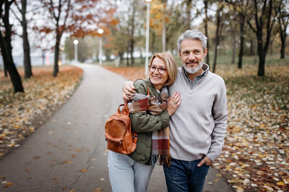 A smiling older couple walks arm in arm along a park path covered in fallen autumn leaves.