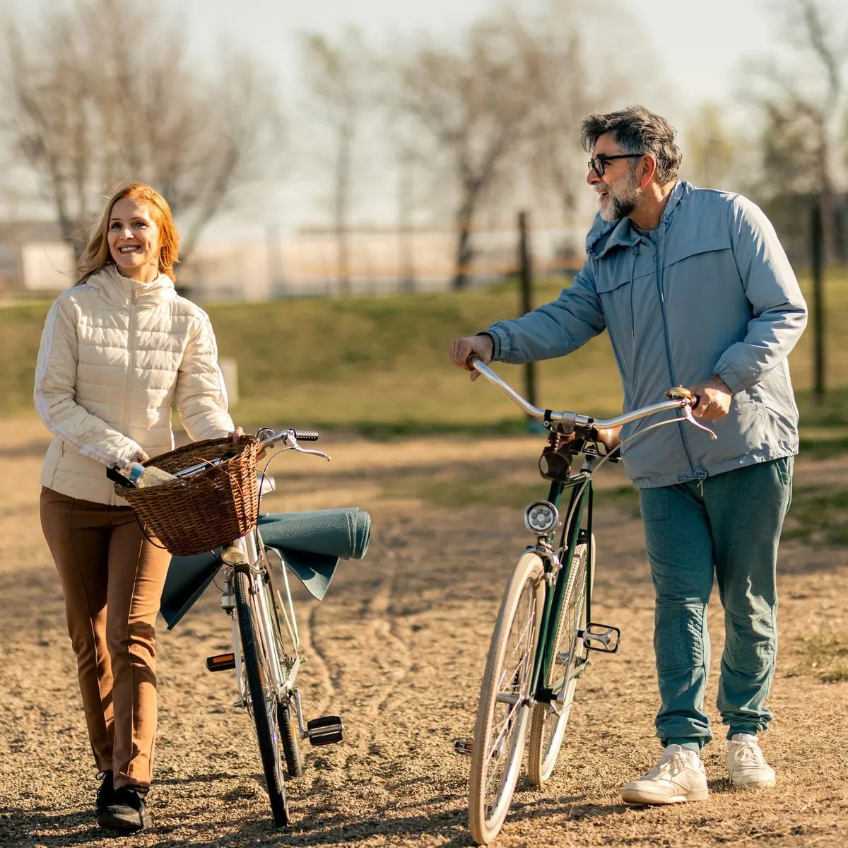 A man and a woman in casual outdoor clothing walking alongside their bicycles on a dirt path.