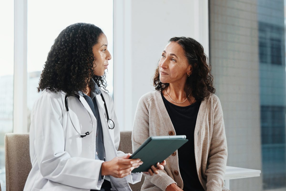 A doctor in a white coat holding a tablet talks to a patient.