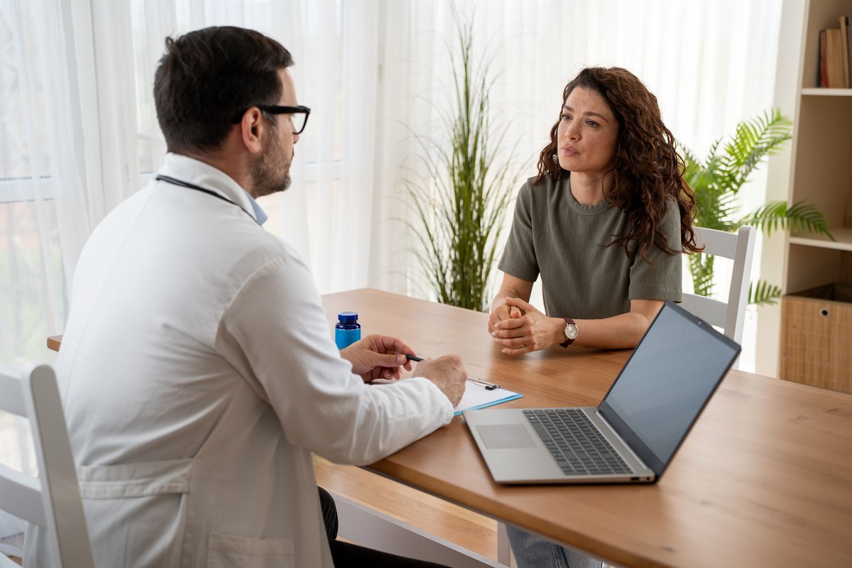 A doctor in a white coat sits at a desk across from a patient during a consultation.