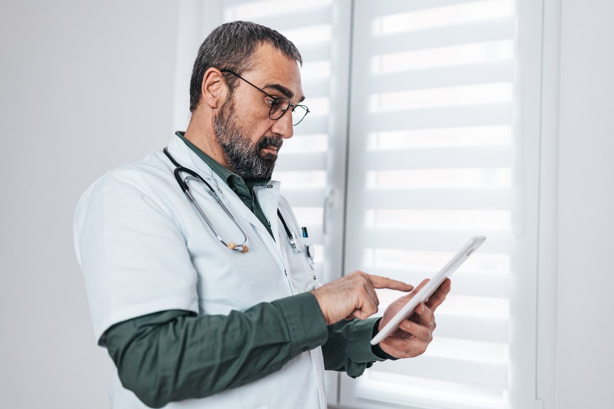 A doctor in a white coat and glasses looking at a tablet.