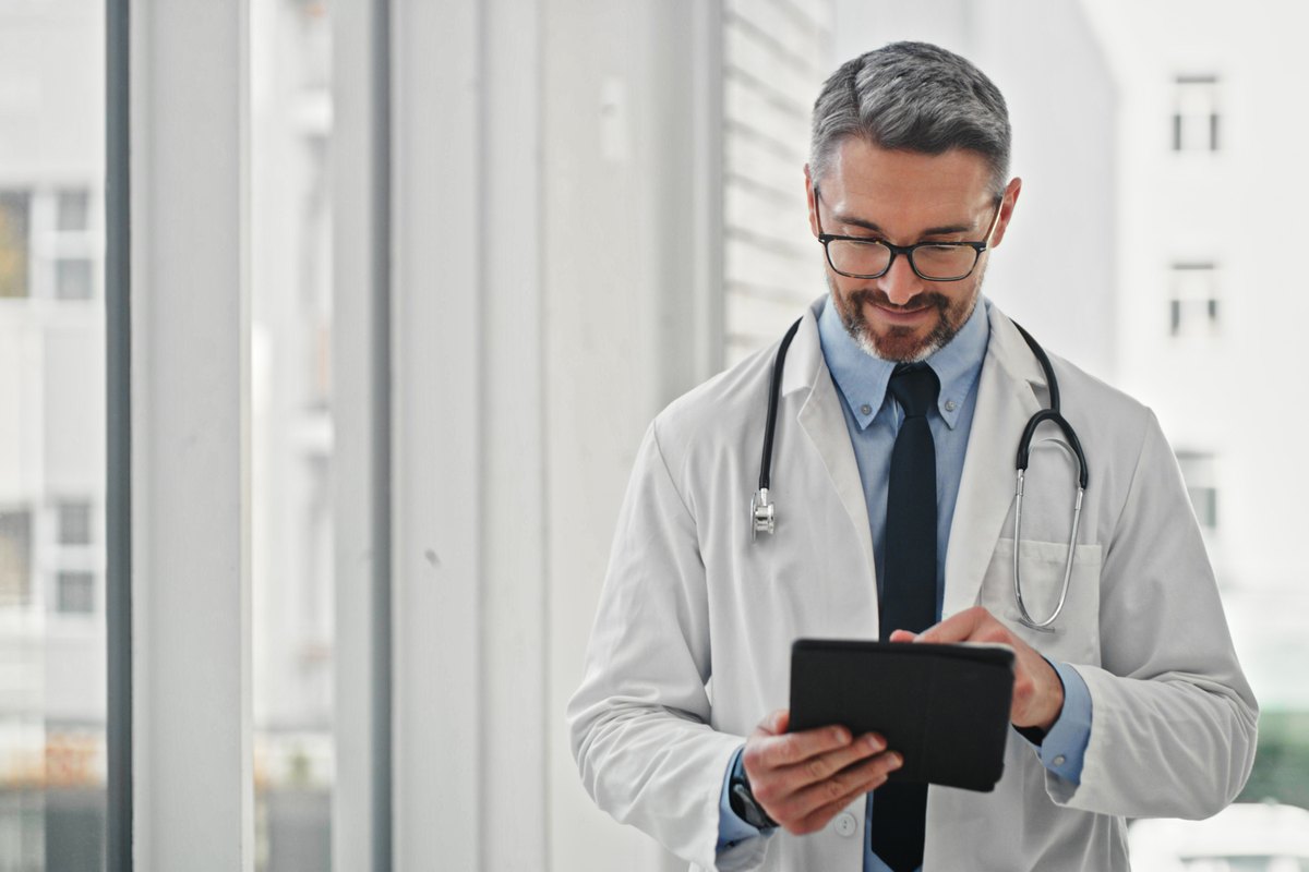 A doctor with a stethoscope around his neck looks down at a tablet computer.