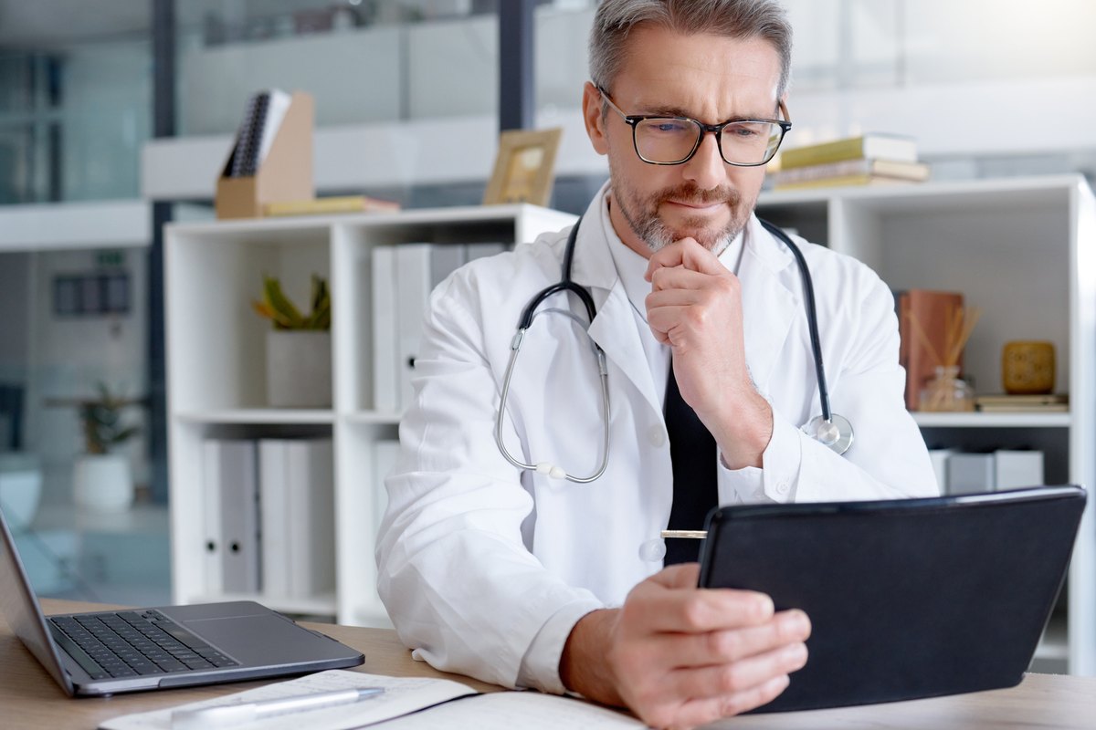 A middle-aged male doctor with glasses and a stethoscope around his neck looks thoughtfully at a tablet while sitting at his desk.