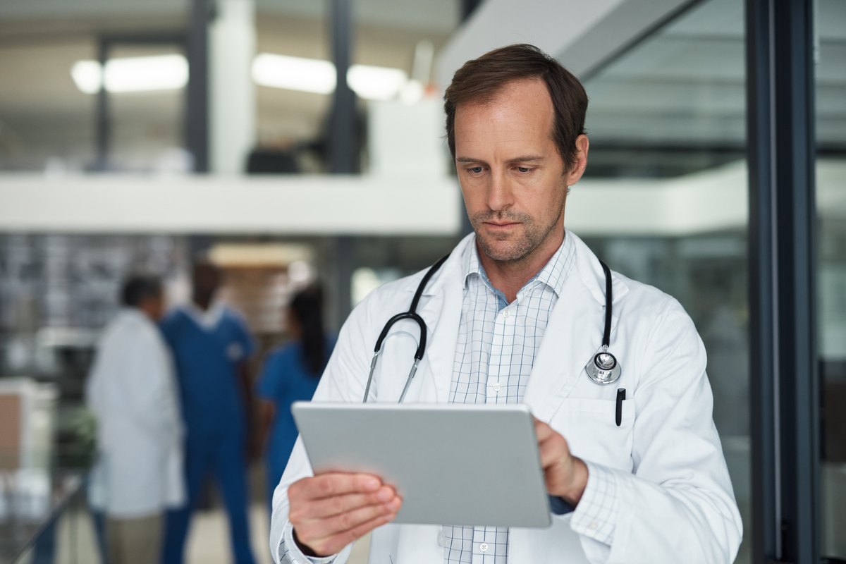 A doctor in a white coat and stethoscope looking at a tablet in a hospital hallway.