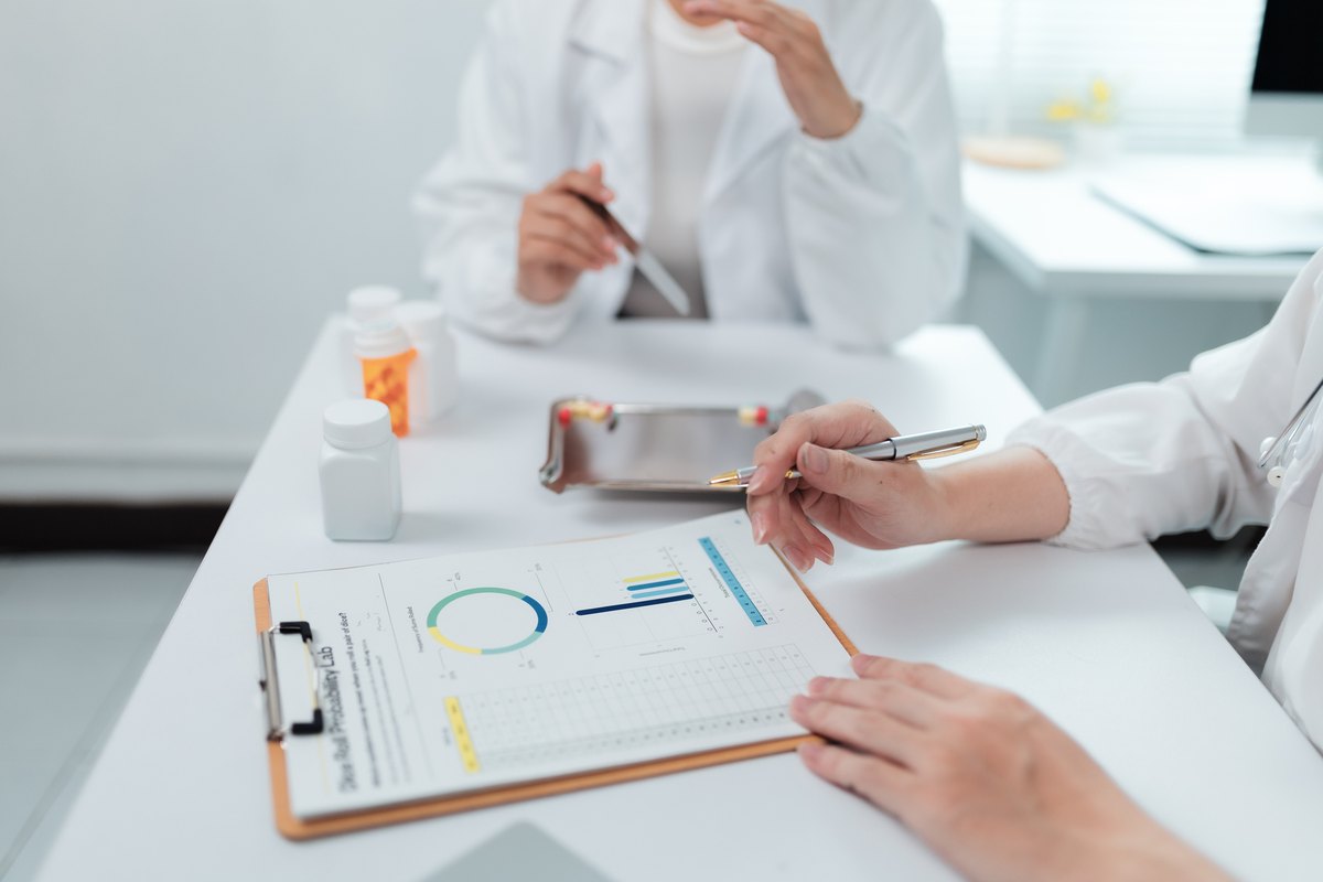 Two healthcare professionals review a clinical report with a pie chart and bar graph on a clipboard at a table with pill bottles.