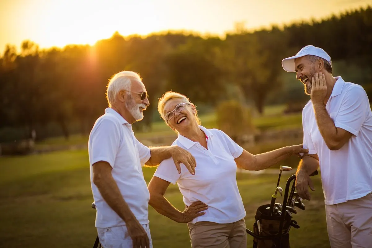 Three friends in white shirts laughing together on a golf course at sunset.