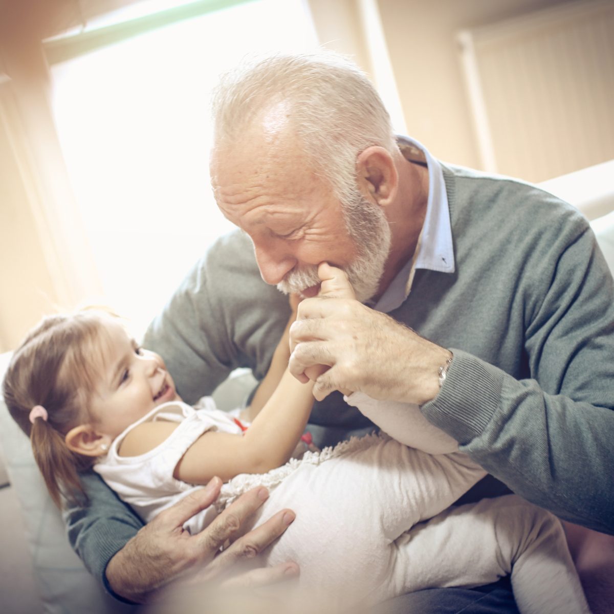 An older man with a grey beard smiles while holding the foot of a laughing toddler sitting on his lap.