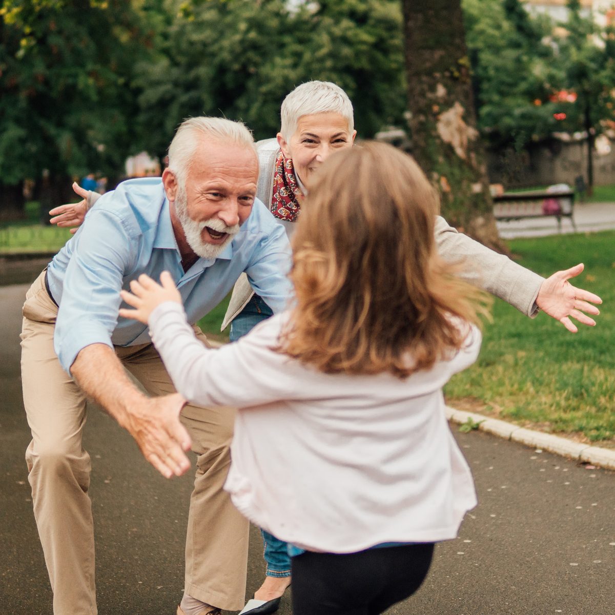 A young girl running with open arms toward her smiling grandparents in a park.