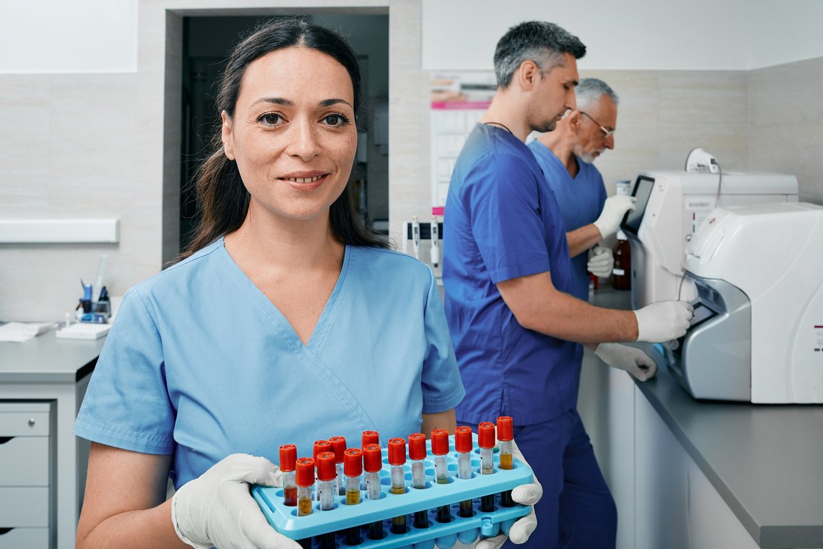 A healthcare worker in blue scrubs holds a rack of test tubes with red caps while colleagues work with laboratory equipment in the background.