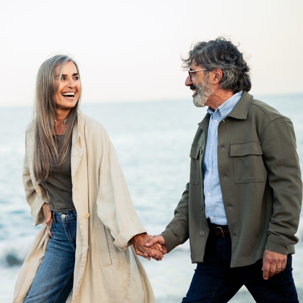 An older couple holding hands and laughing while walking together along the beach.