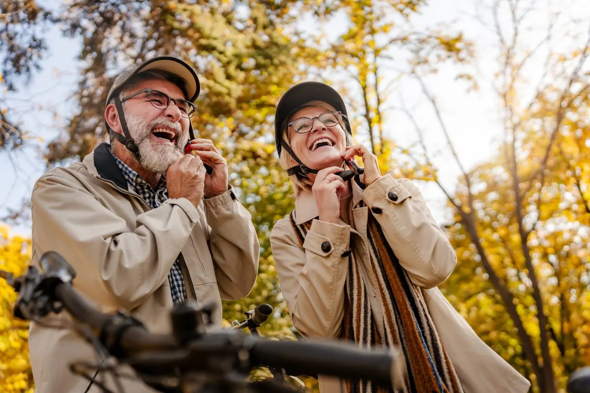 An older man and woman laughing as they adjust their bicycle helmets outdoors during autumn.