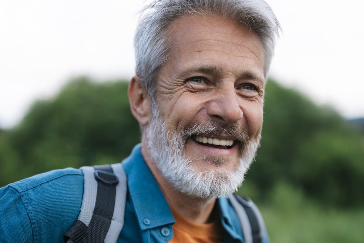 A smiling man with a grey beard wearing a backpack outdoors.