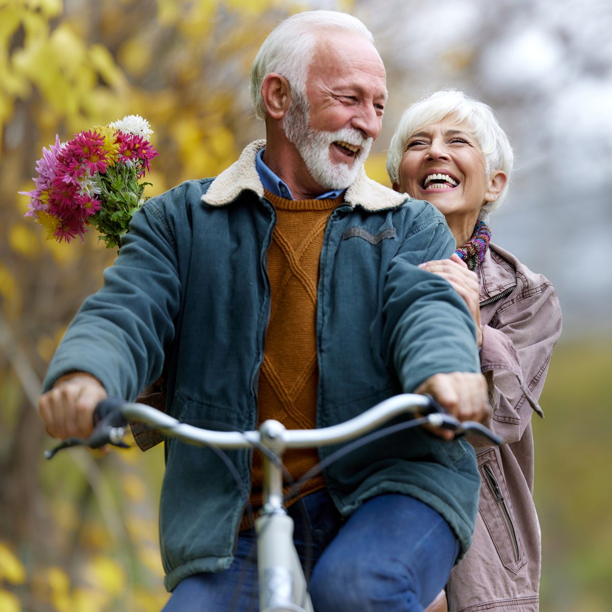 An elderly couple laughing while riding a bicycle together, with the man holding a bouquet of colorful flowers.