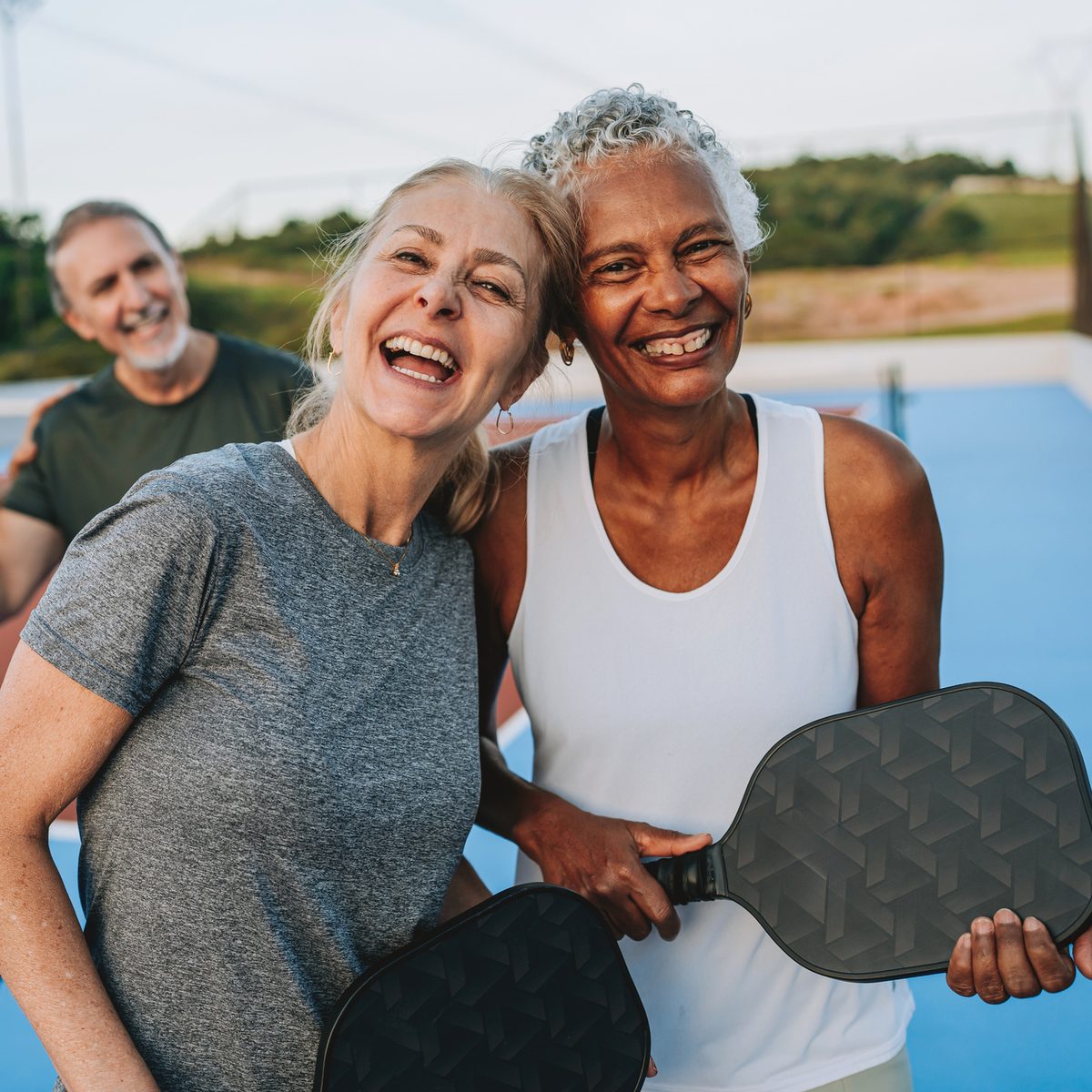 Two women smiling and holding pickleball paddles on a court, with a man smiling in the background.