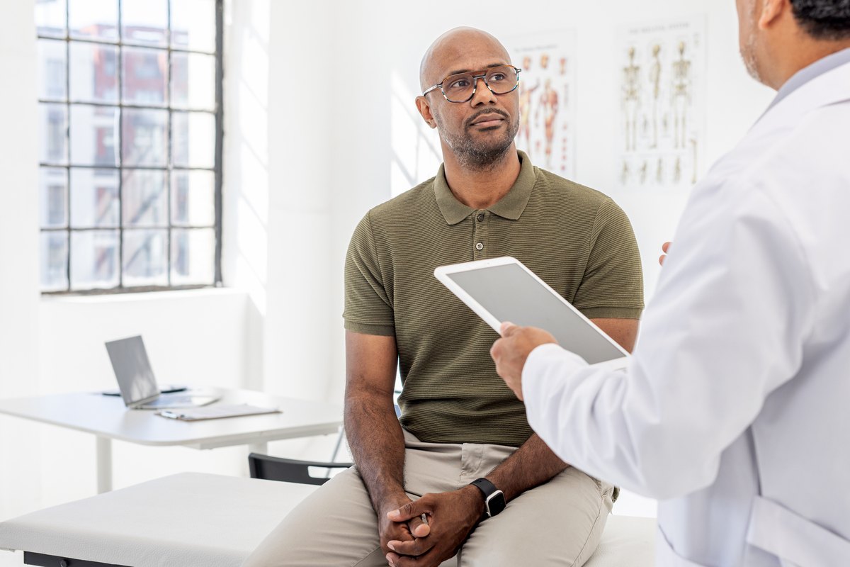 A doctor holding a tablet speaks with a patient sitting on an exam table.