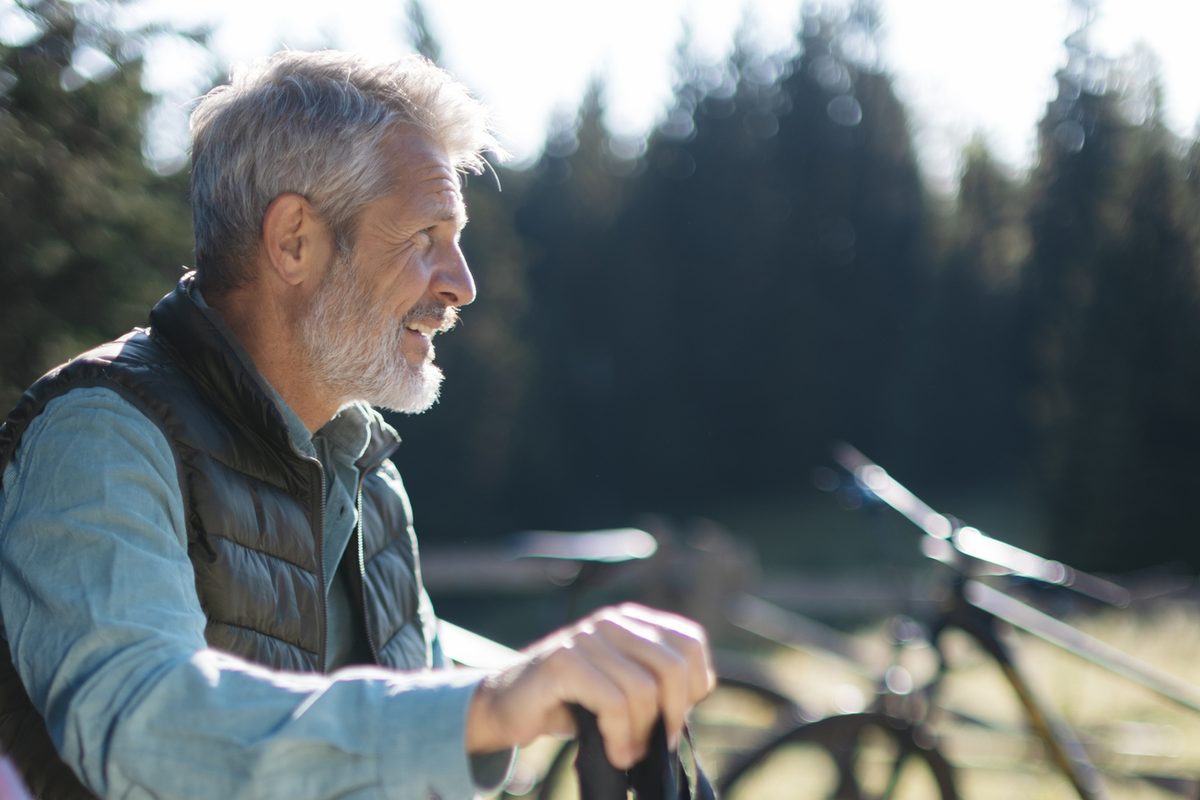 A smiling, gray-haired man in a light shirt and puffer vest sits outdoors with a bicycle visible in the background.