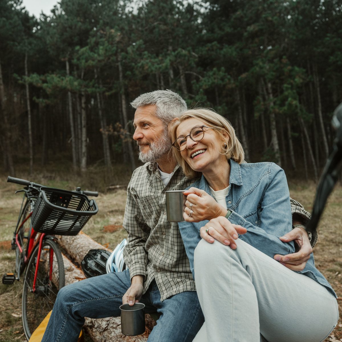 An older couple sitting on a fallen log in the woods, holding mugs and smiling.