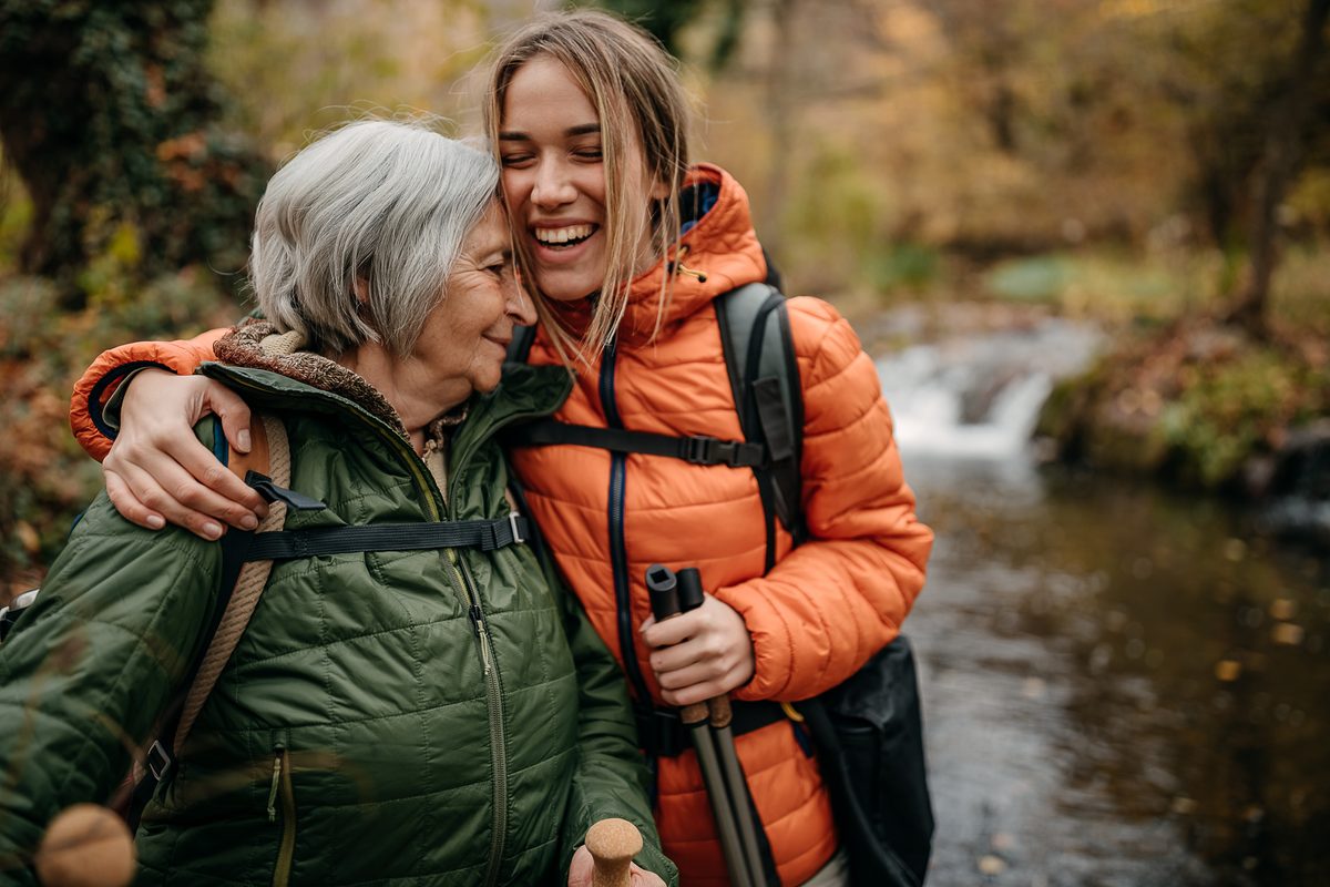 An older woman and a young woman in hiking gear embrace while standing beside a stream.