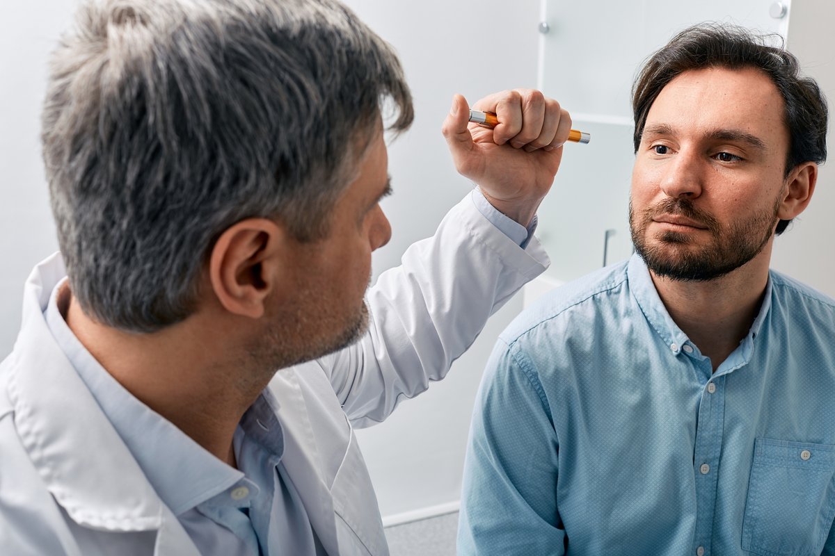 A doctor in a white coat uses a penlight to examine a male patients eye.