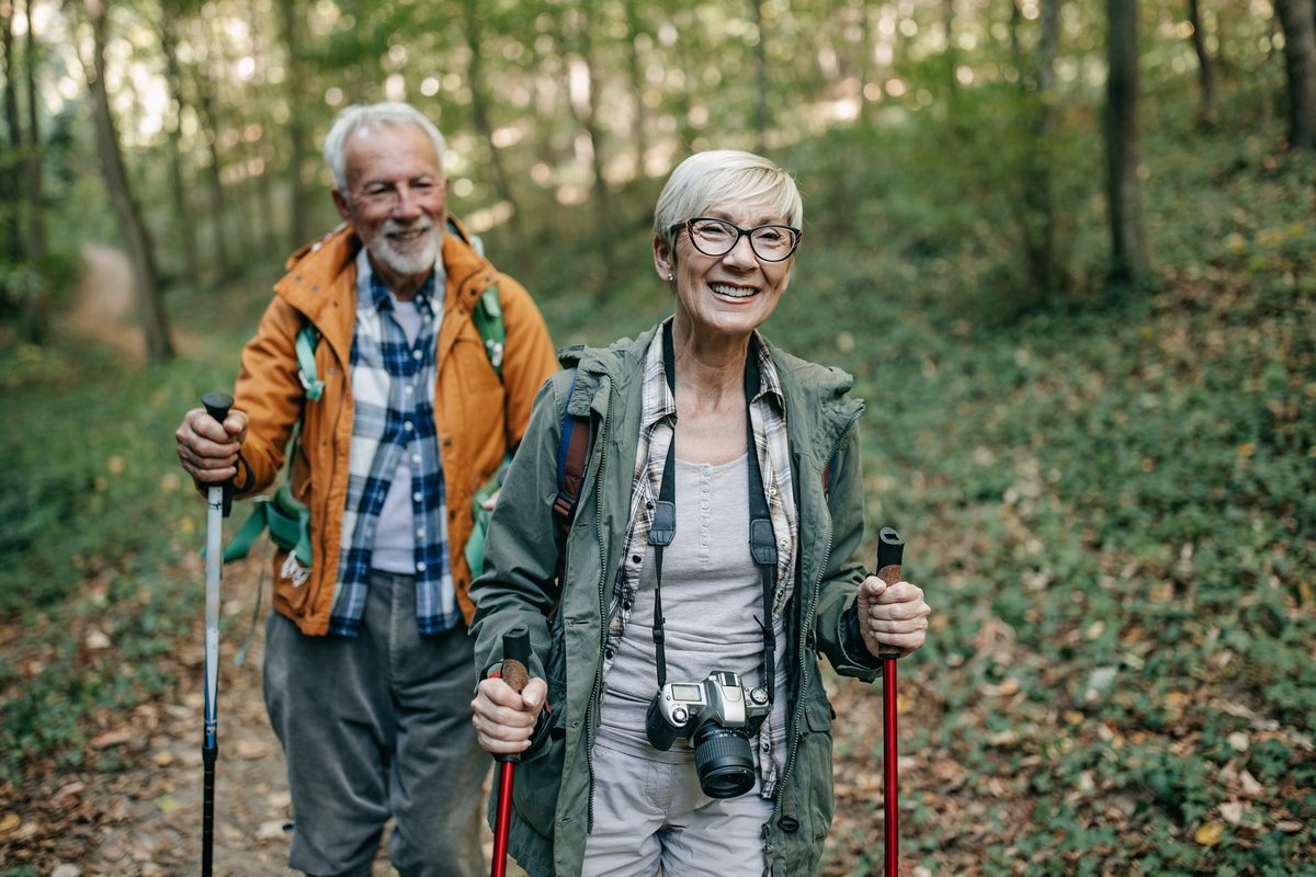 An older couple with trekking poles smile while hiking on a forest trail.