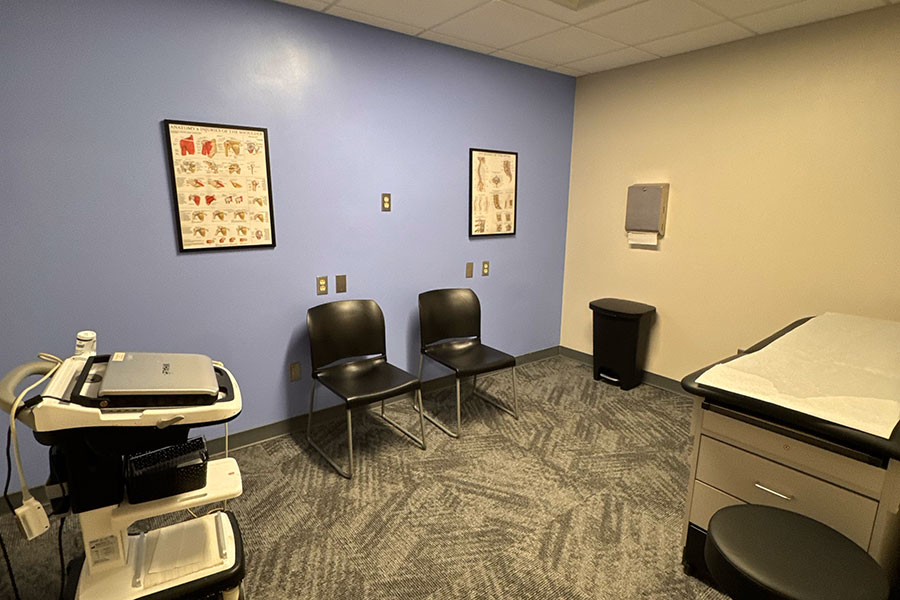 A Grand Rapids office exam room featuring two chairs, a mobile laptop cart, a wall-mounted paper dispenser, a trash can, and an exam table with a stool.