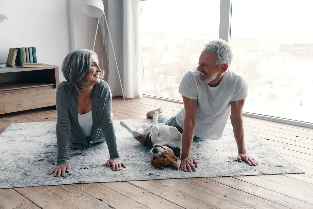 An older couple smiles at each other while doing yoga on a rug at home, with a beagle lying on its back between them.