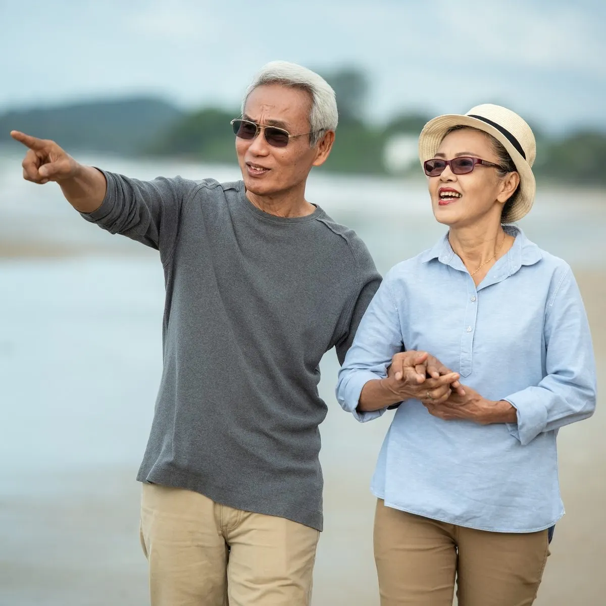 An older couple walks on a beach, with the man pointing toward the distance while holding the womans hand.