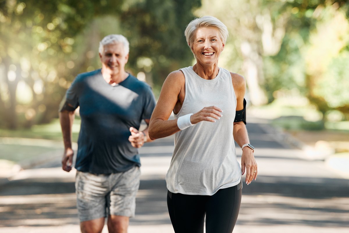 A smiling older woman runs on a paved path with a man jogging behind her.