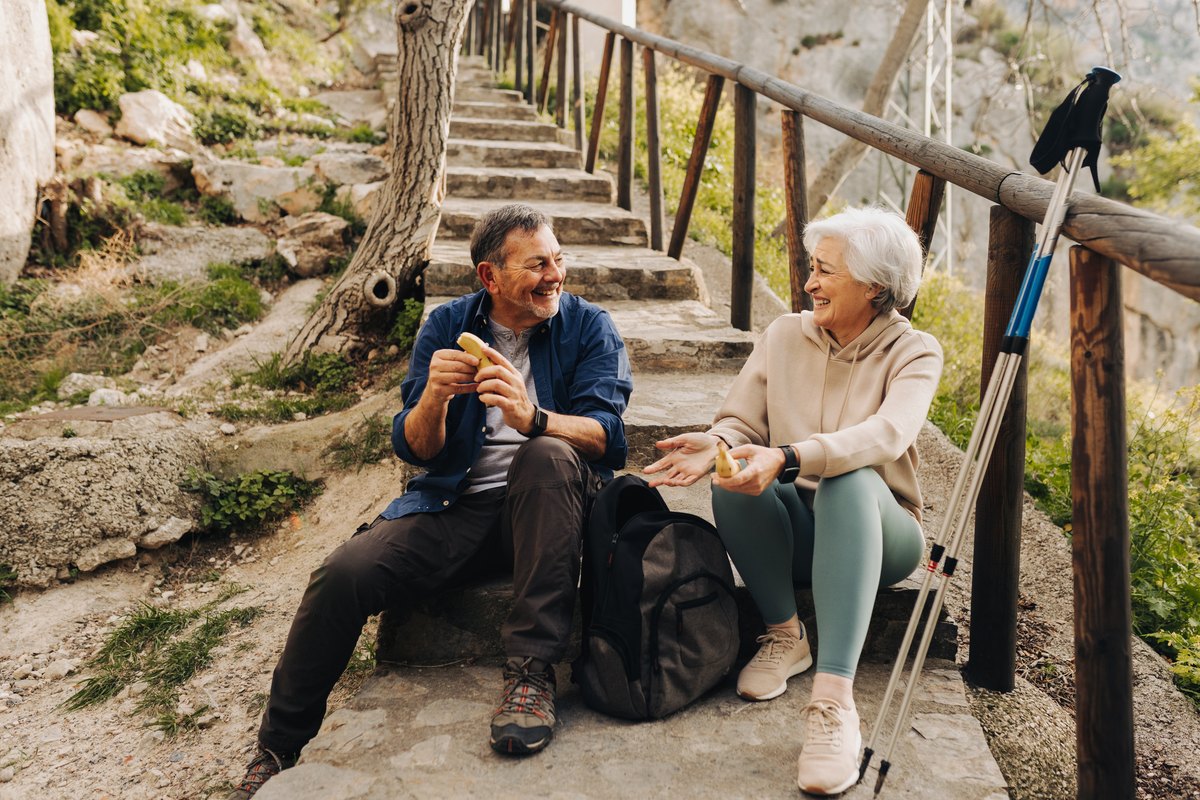 An older couple sitting on stone steps during a hike, smiling and eating snacks.