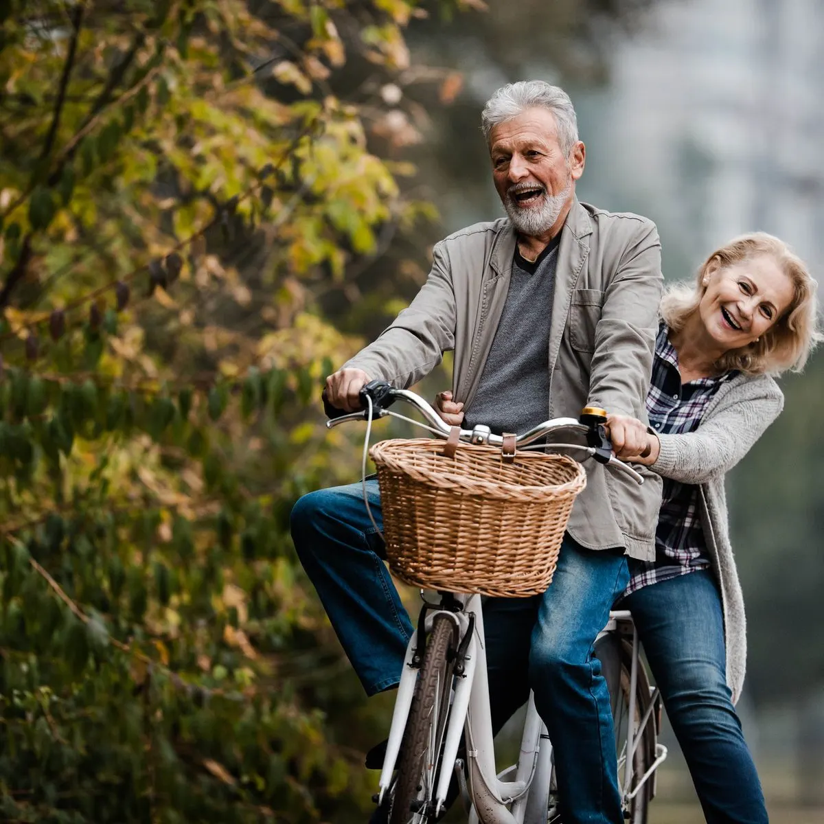An older couple laughs while riding a bicycle together outdoors.