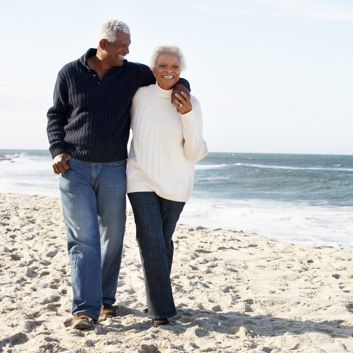 An older couple walks arm in arm along a sandy beach.