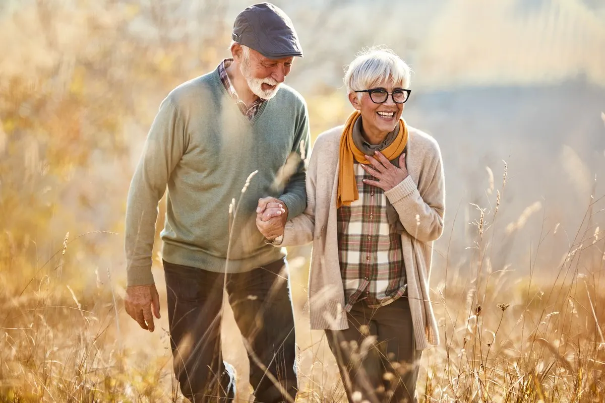 An older couple holding hands and smiling while walking through a field of tall grass.