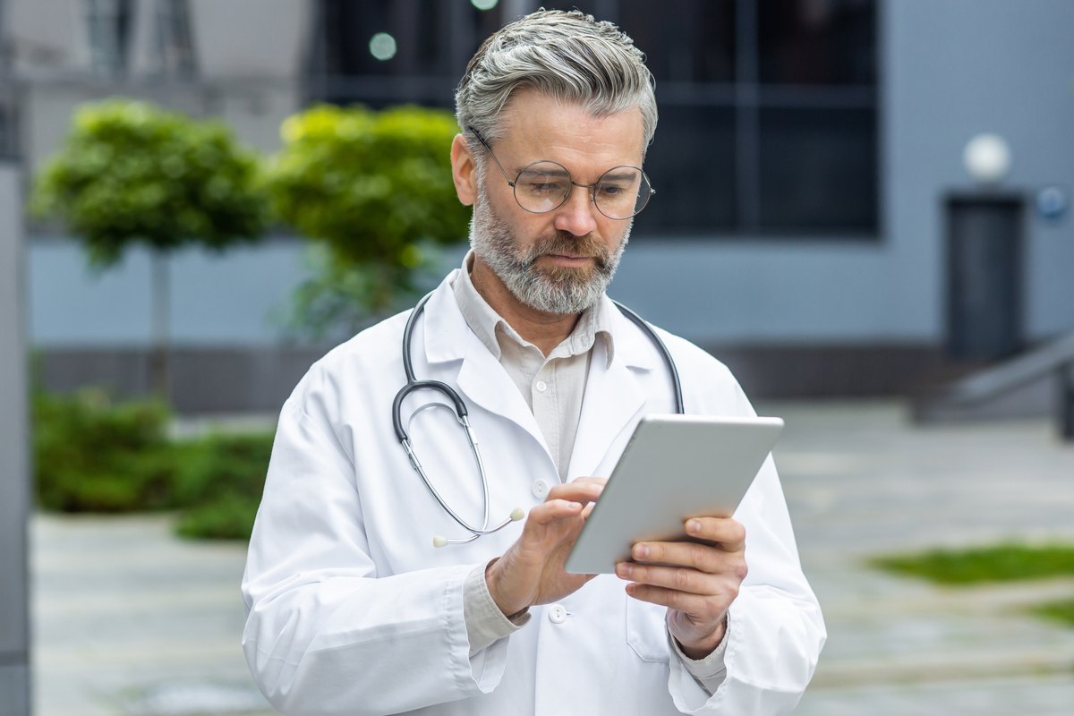 A middle-aged doctor with a beard and glasses in a white lab coat uses a tablet.