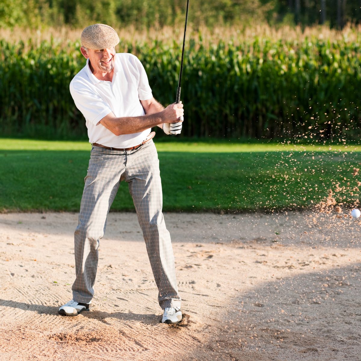 An older man in a flat cap and golf attire hits a golf ball out of a sand bunker.